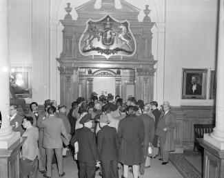 Une foule d’hommes se tient devant une porte ouvragée. Juste au-dessus des têtes, le nombre « 24 » est visible.