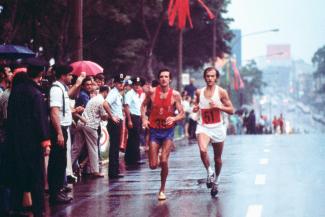 Photographie de deux athlètes courant dans la rue sous la pluie, avec une foule alignée sur le côté.